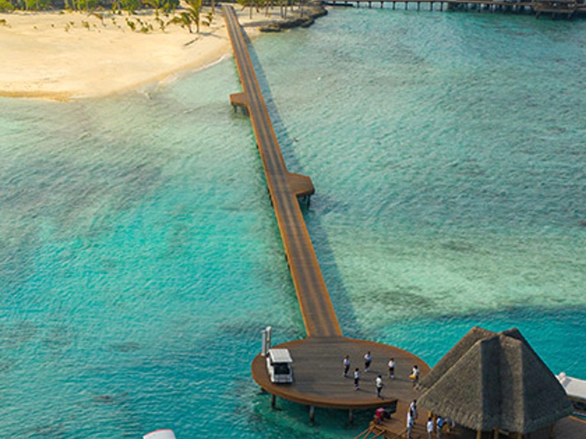 Aerial view of a long timber jetty crossing shallow turquoise reef water to the island.