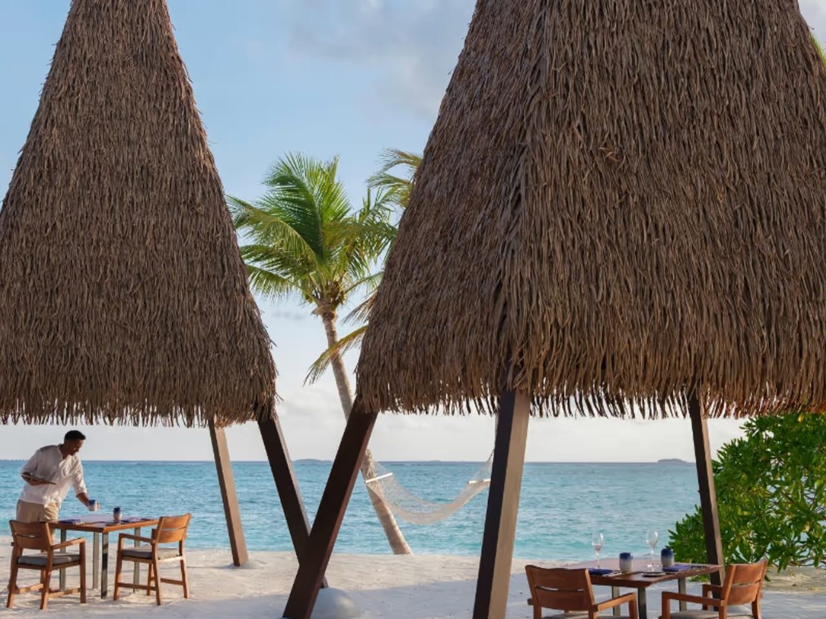 Tall thatched A-frame cabanas over dining tables on the white-sand beach, with palms and the turquoise sea behind.
