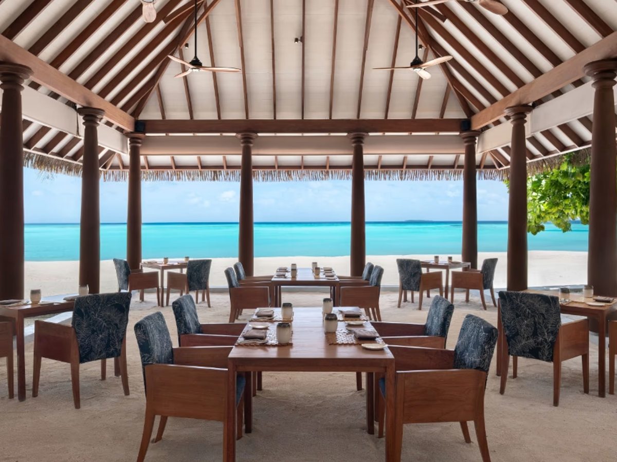 An open-sided restaurant with wooden tables and chairs under a high timber roof, looking out over the pool and lagoon.