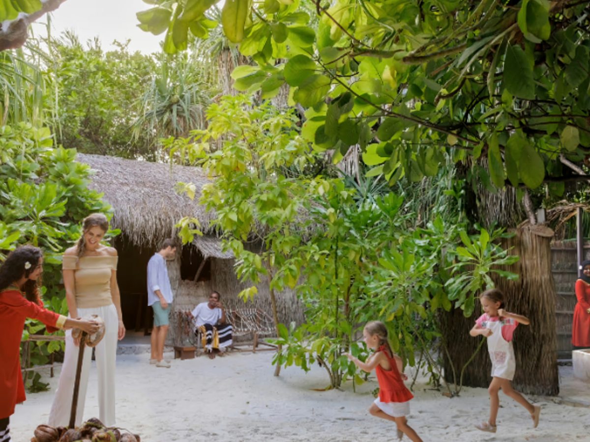 A family walking a sandy path between dense tropical planting and thatched villas on the island.