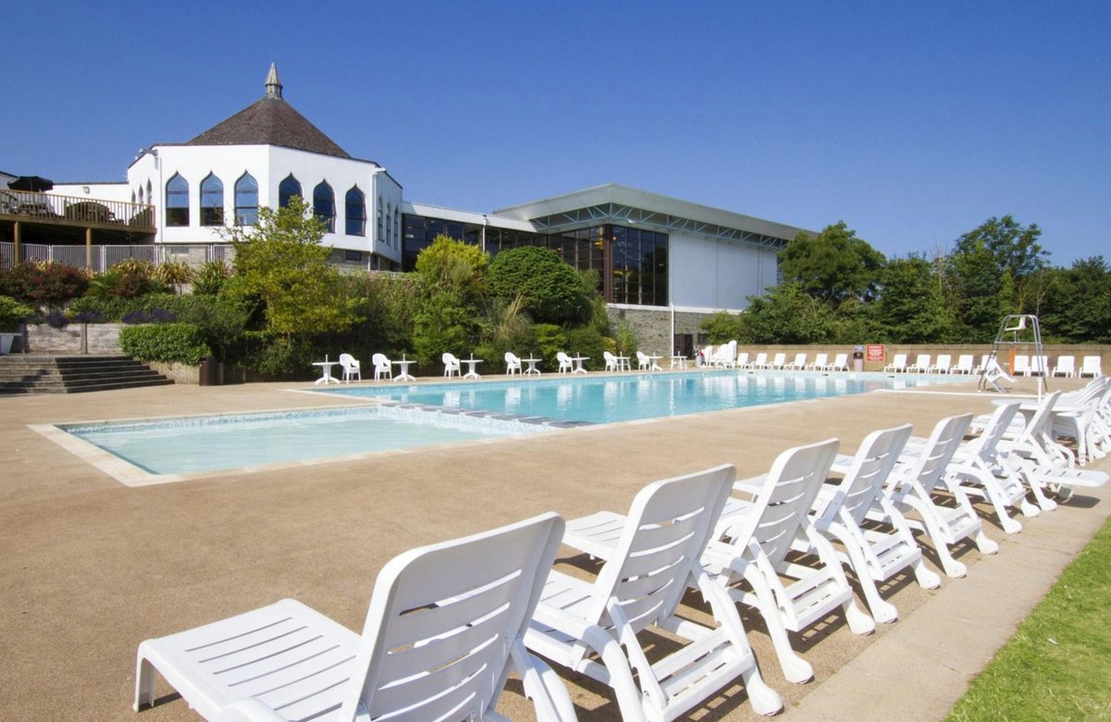 The outdoor pool at Hoburne Devon Bay, lined with white loungers below the octagonal leisure complex under a clear blue sky.