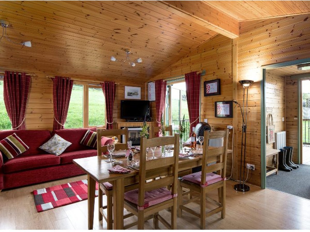 Open-plan log-cabin living room with a red sofa, a wooden dining table set for a meal, pine-clad walls and ceiling, and countryside through the windows.
