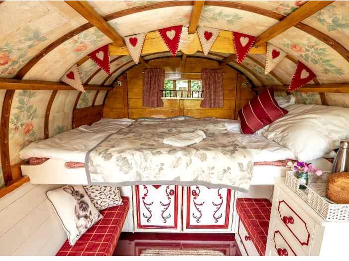 The painted interior of a bow-top gypsy caravan with a raised double bed under a floral-lined arched roof, heart bunting, cushioned bench seats and a small dresser.
