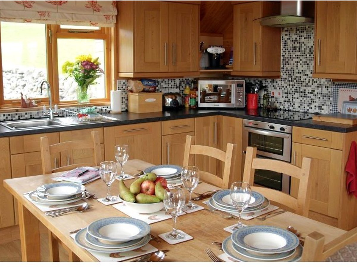 A fitted wooden log-cabin kitchen with an oven, hob, microwave and mosaic-tiled splashback, and a table laid with crockery, wine glasses and a bowl of fruit.