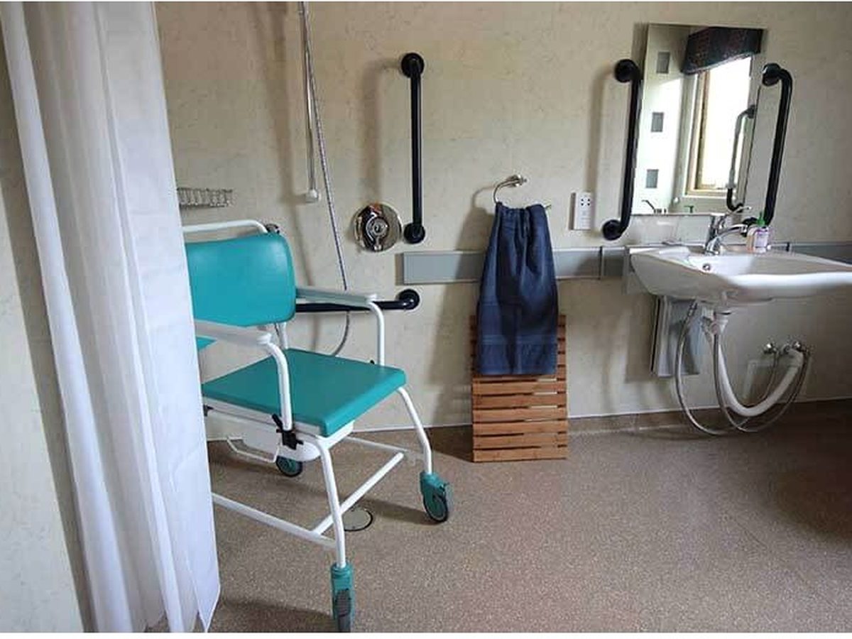 A wet room with a wheeled shower chair, wall grab rails, a wall-hung basin with mirrors and a level floor, with a window to one side.