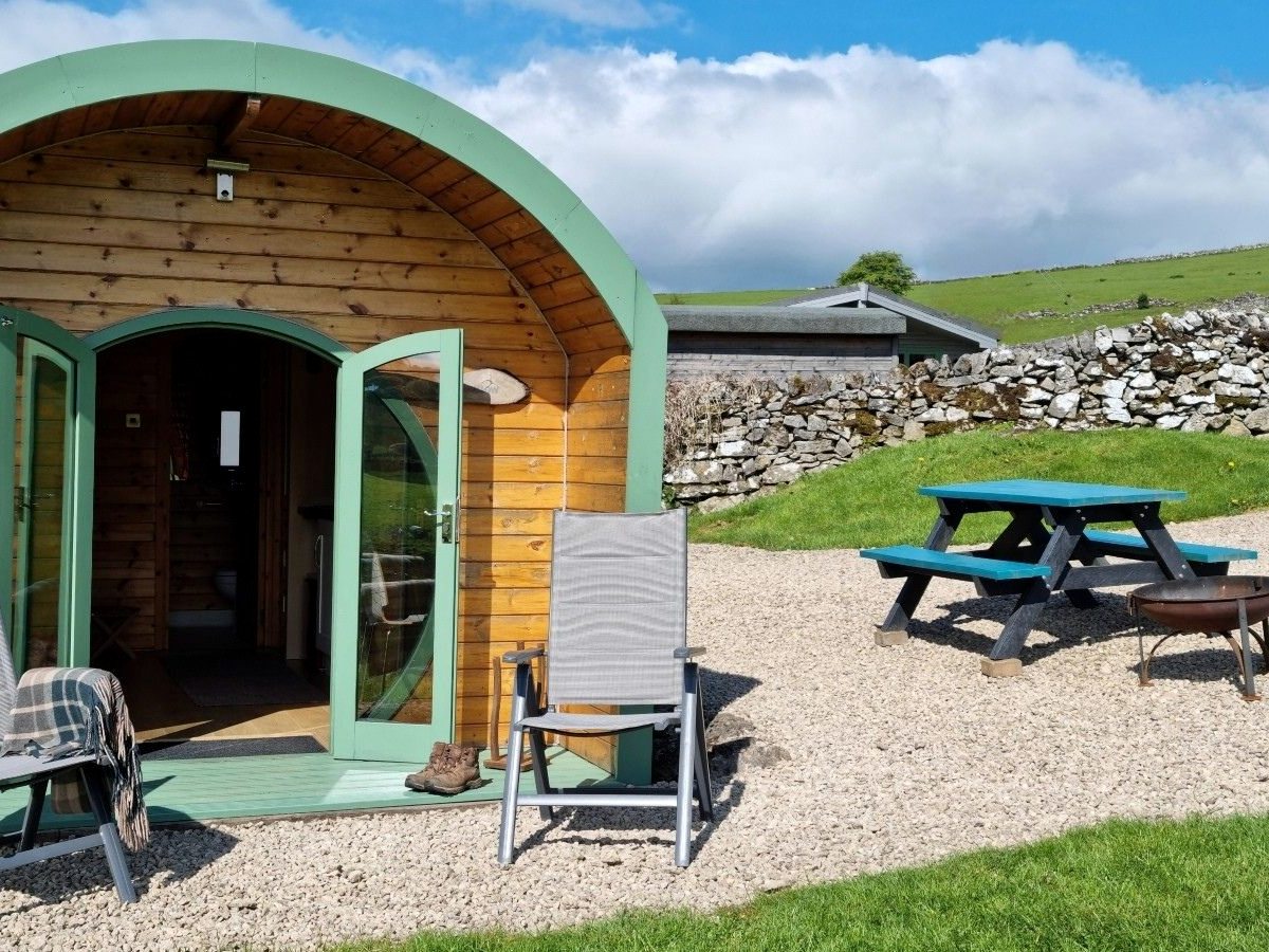 A timber glamping pod with a curved roof and green-framed doors, a picnic bench and firepit on gravel beside a dry-stone wall, with a cabin and green hills behind.