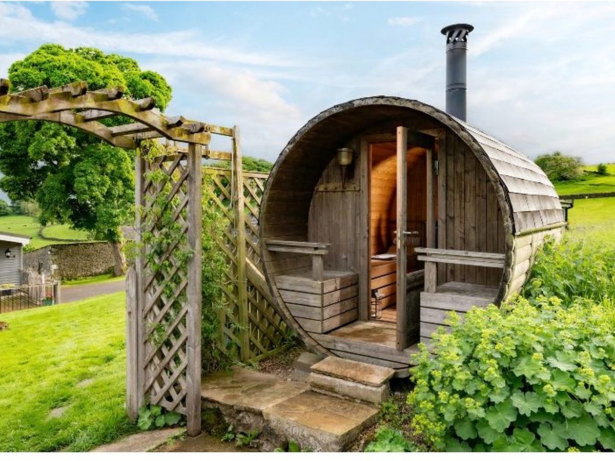 A wooden barrel-shaped sauna with a metal flue and a small round window, set in a garden beside a timber pergola arch, with green fields behind.