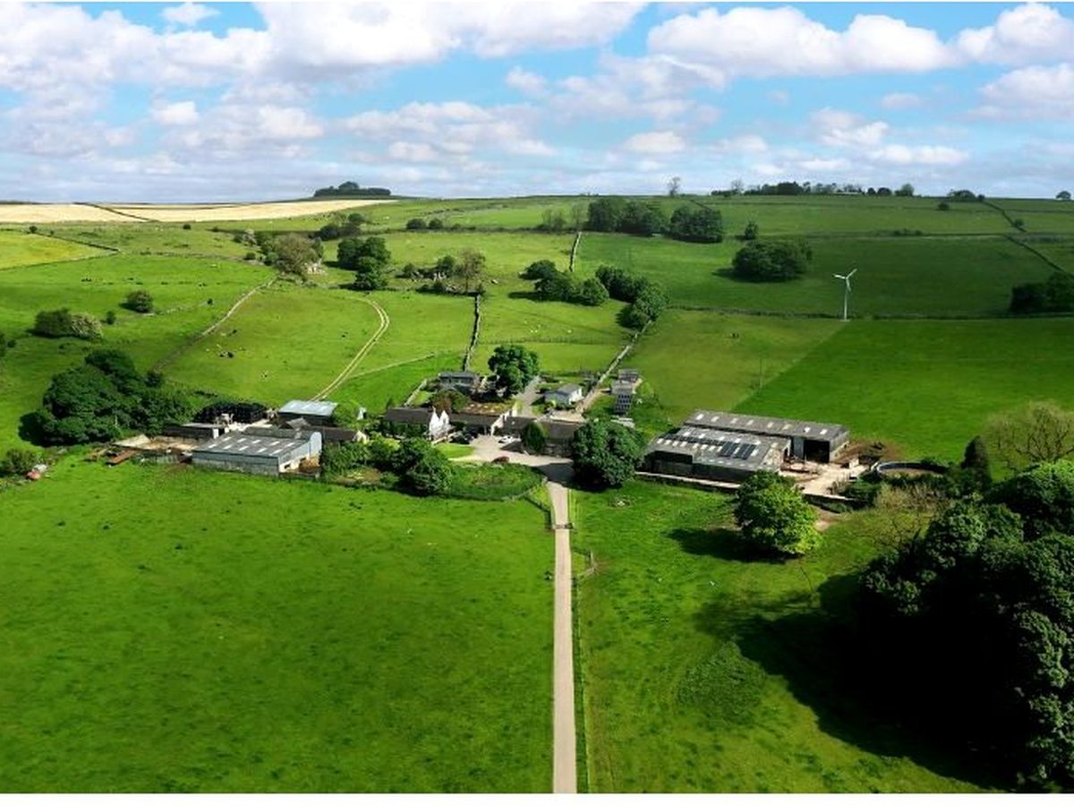 Aerial view of the farm's buildings and barns set among a patchwork of green fields bounded by dry-stone walls, with a wind turbine and open countryside beyond.