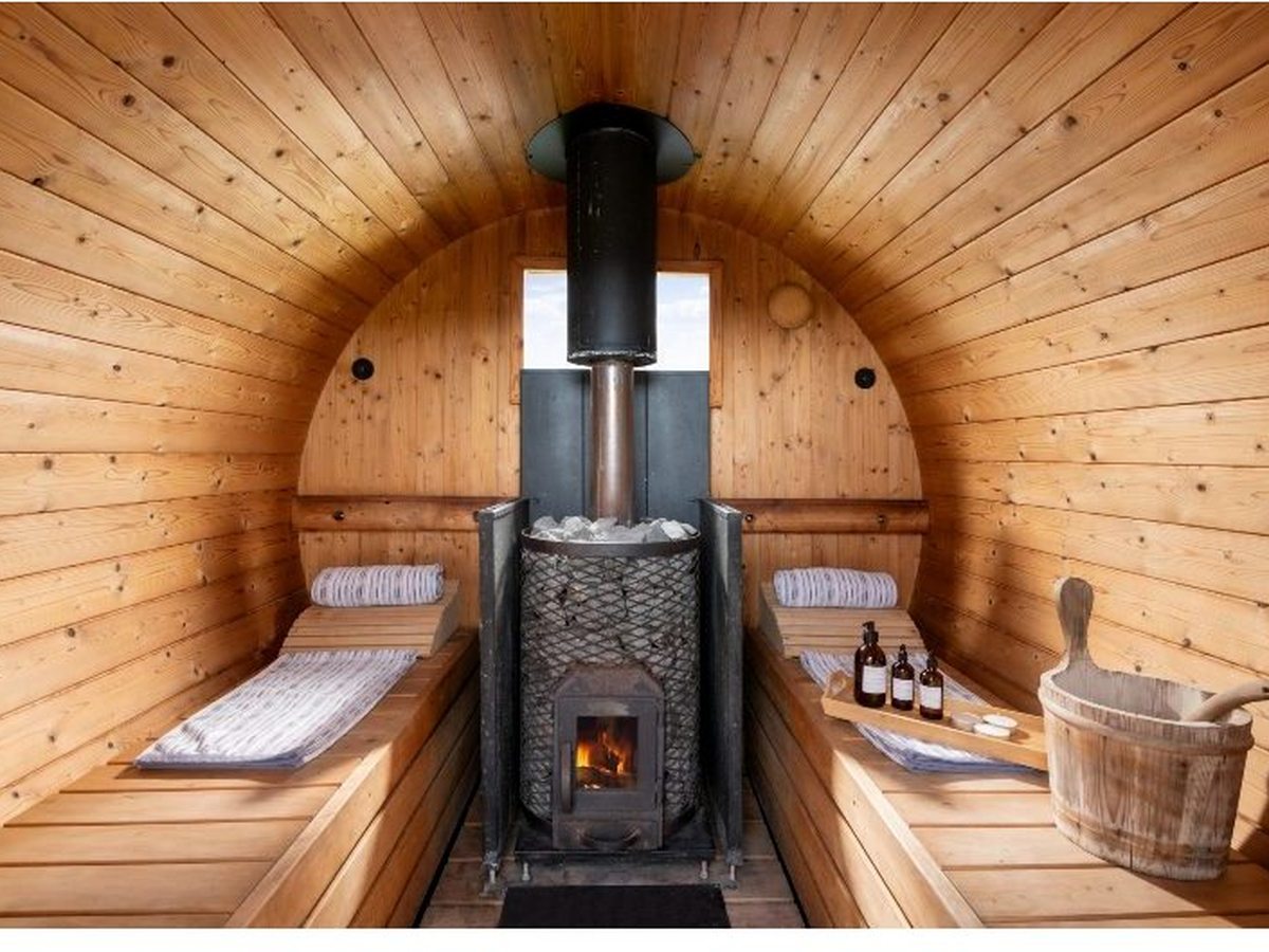 The pine interior of a barrel sauna with facing bench seats, a central wood-fired stove and flue, and a wooden water bucket and ladle.