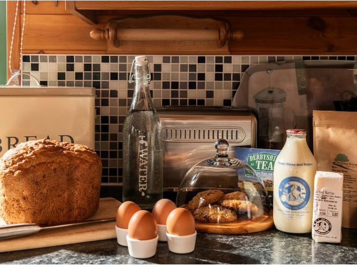 A kitchen worktop with a fresh loaf by a bread bin, a bottle of water, boiled eggs in egg cups, home-baked biscuits under a glass dome, a bottle of milk, salted butter, Derbyshire tea and Hoe Grange coffee.