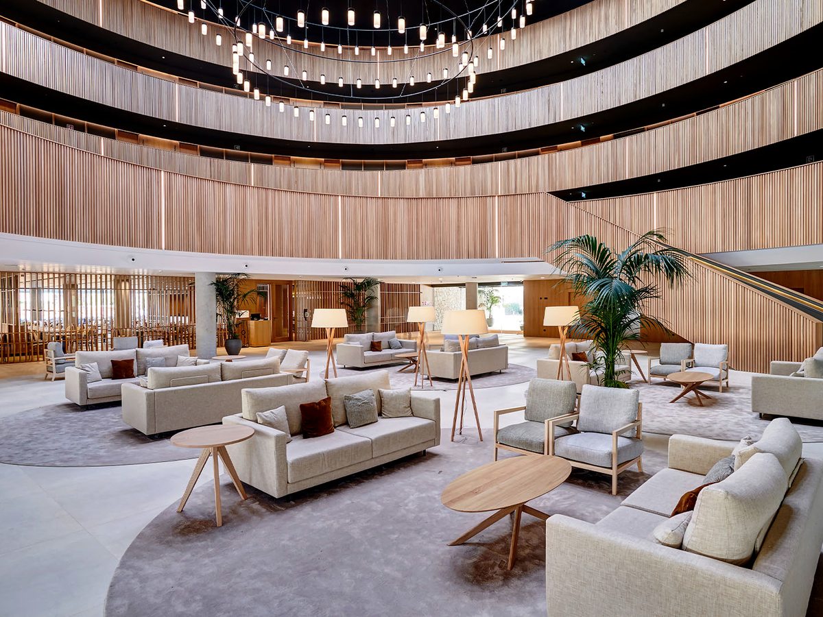 Double-height circular lobby atrium with curved timber-slat walls, pale sofas and armchairs, wooden coffee tables and cluster pendant lights over a polished floor.