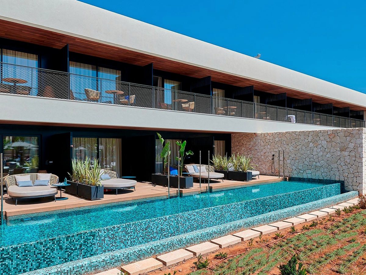 Ground-floor room with a wooden deck and cushioned daybeds opening onto a long mosaic-tiled swim-up channel, with a stone wall and balconied block above.