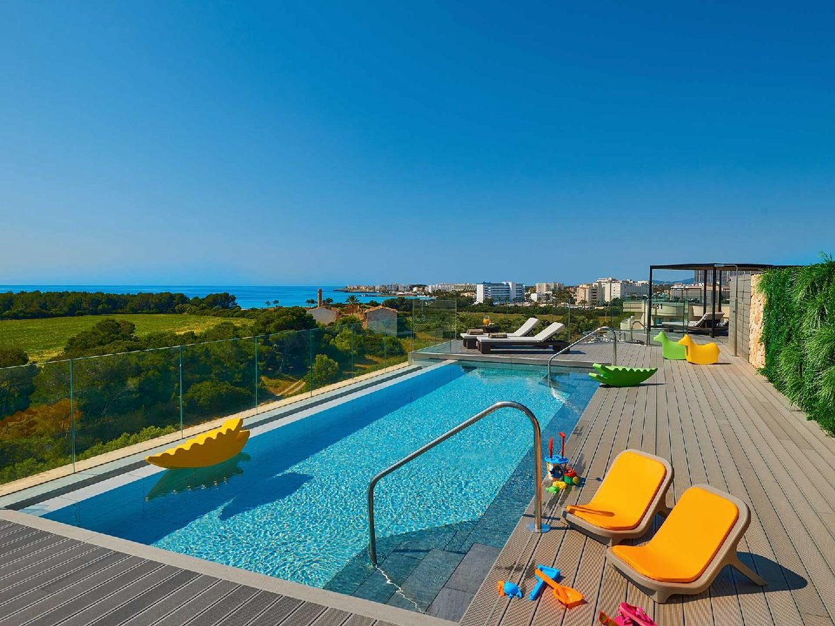 Elevated deck pool with a glass balustrade and colourful children's rocker toys, looking out over green countryside and a coastal town to the sea.