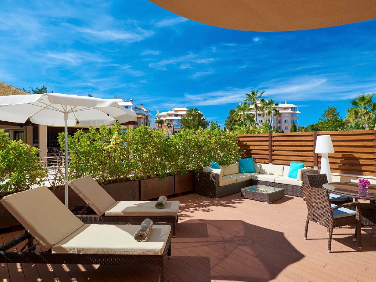 Private roof terrace with cushioned sun loungers, a corner sofa and a dining table under a shade sail, looking over the resort's white apartment blocks and palms.