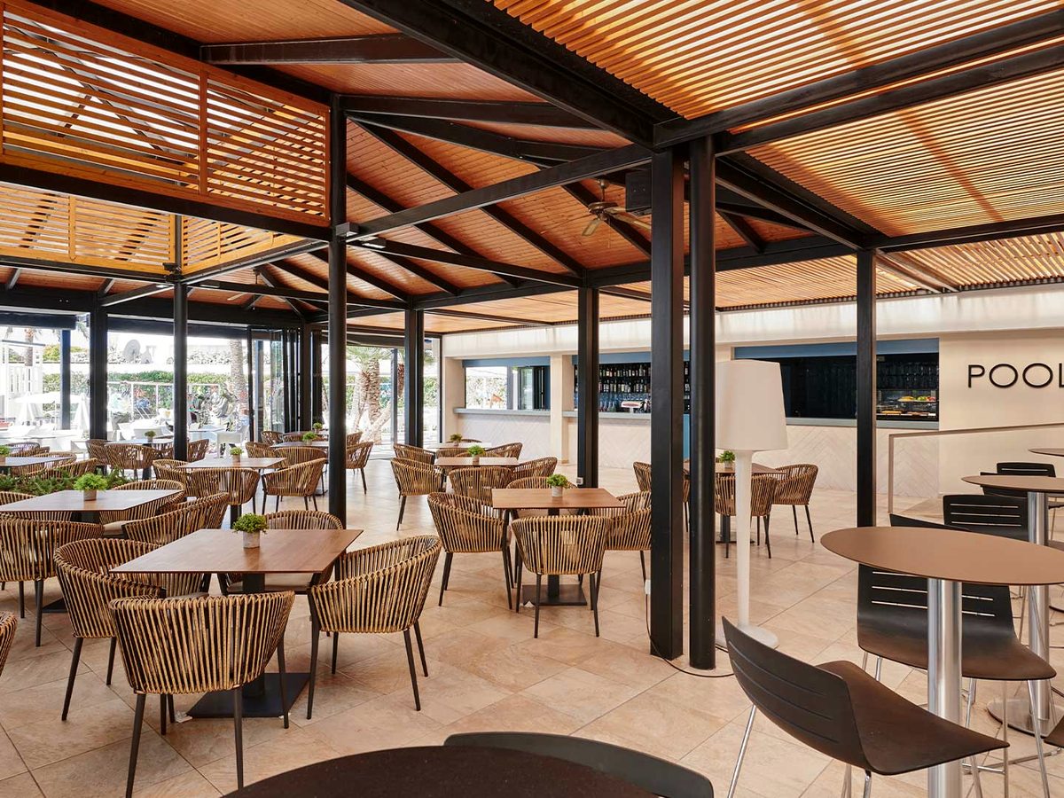 Covered poolside bar with a timber-slat pergola roof, rattan tub chairs at wooden tables and a bar counter, open on one side to the pool.