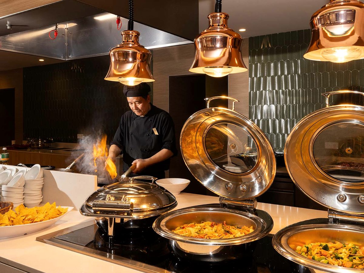 Buffet live-cooking station with a chef flambeing at a hob under copper heat lamps, beside stainless chafing dishes of pasta and vegetables.