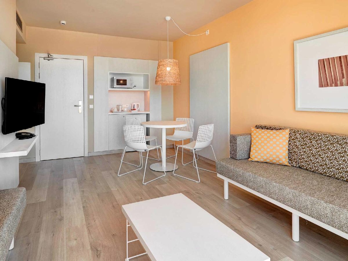Apartment living area with a light wood floor, an apricot accent wall, a fabric sofa bed, a white table with wire chairs and a kitchenette with microwave and kettle.