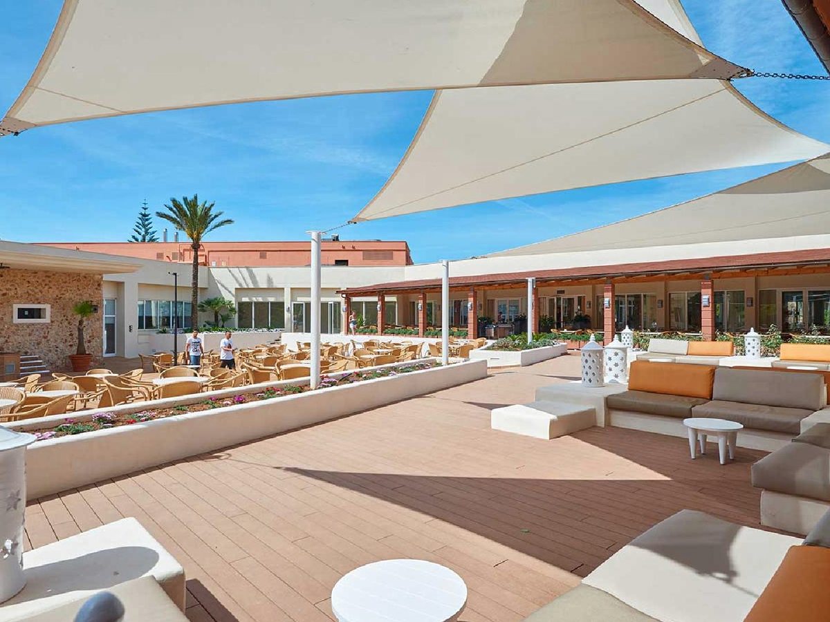 Raised timber deck under white shade sails with low cushioned sofas and rattan chairs, ringed by the resort's ground-floor buildings and a palm tree.