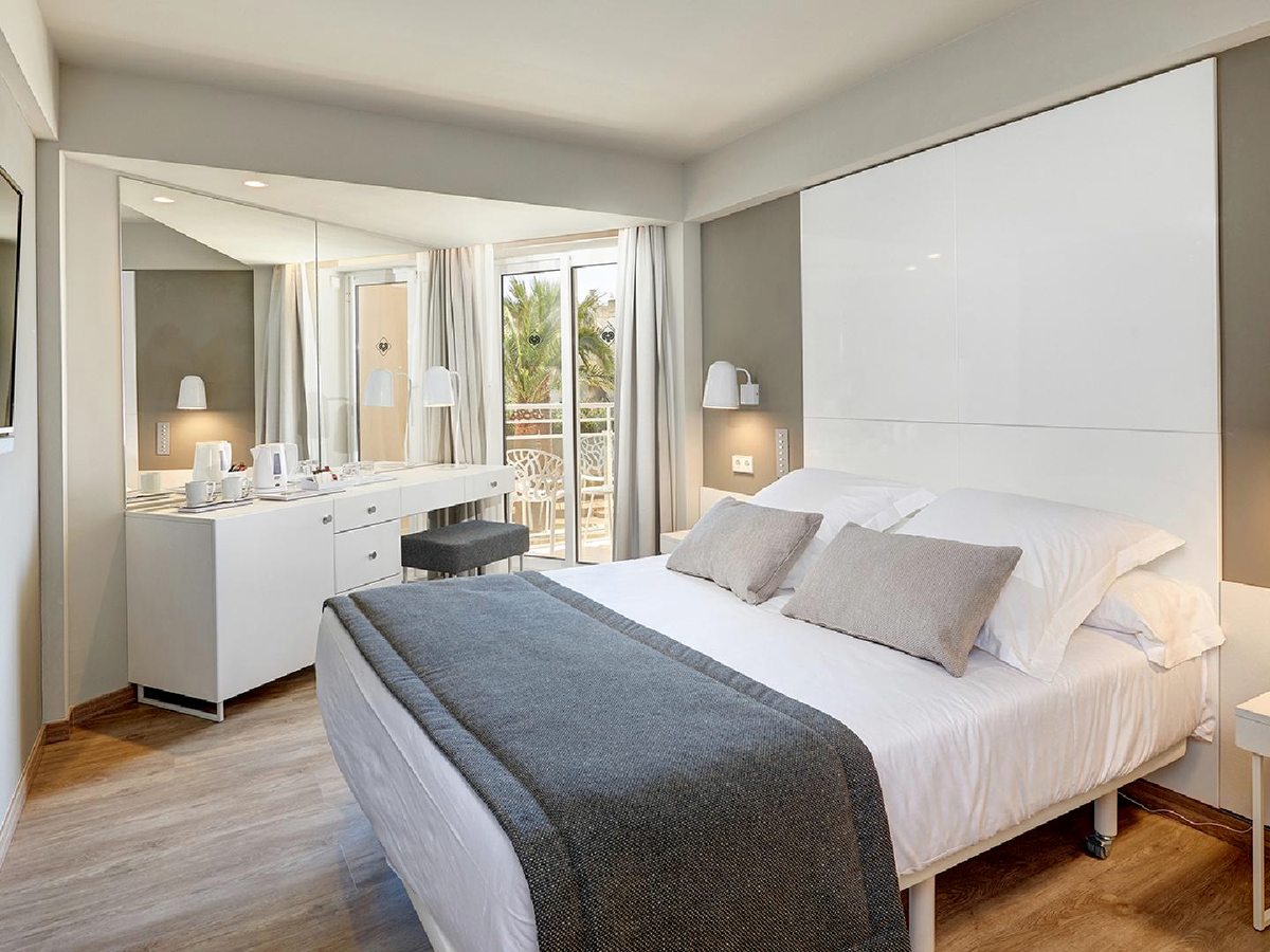 Refurbished bedroom in grey and white with a double bed, a white vanity with a kettle and tea tray, and a curtained balcony with palms beyond.