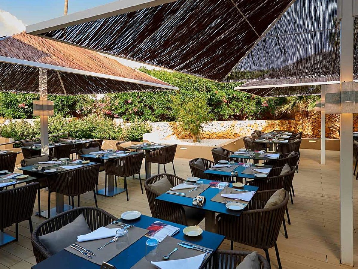 Outdoor restaurant terrace set for dinner with blue-topped tables and rattan chairs under reed parasol shades, beside a dry-stone wall and flowering shrubs.