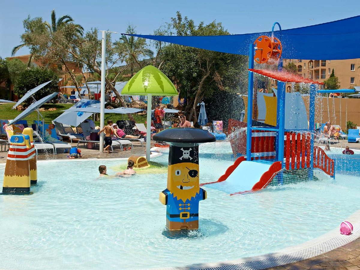 Family splash park with a pirate-themed water-play tower, slides, tipping mechanisms and a green mushroom fountain, with children playing in the shallow water.