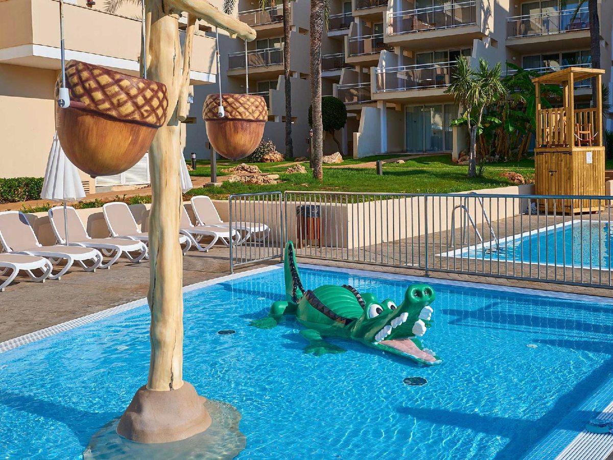 Shallow children's pool with a green crocodile figure and two acorn tipping-buckets on a post, fenced off beside the resort's apartment block.