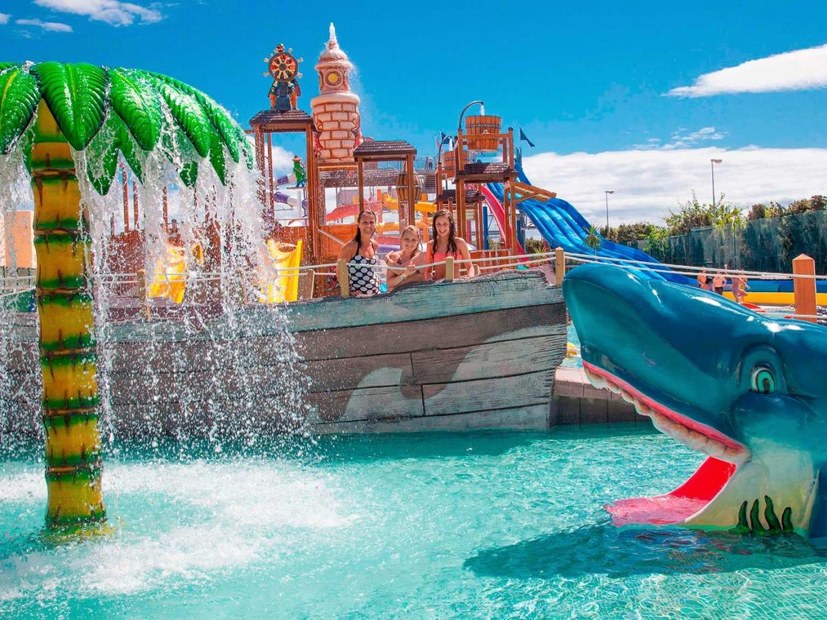 A pirate-ship splash structure with a whale-shaped slide and a palm-tree fountain in a shallow pool, with children playing in the water.