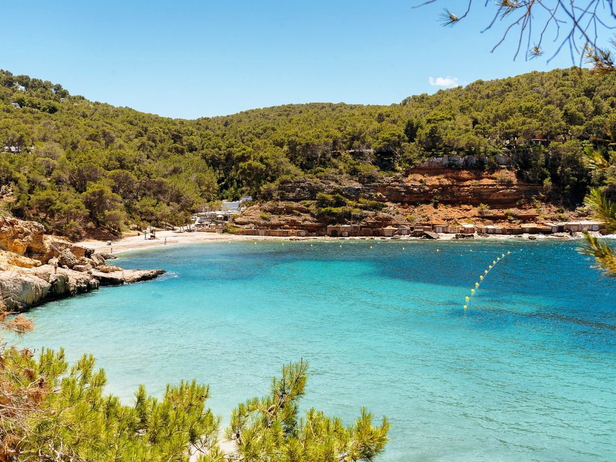 A sheltered cove of turquoise water backed by pine-covered hills and low red-rock cliffs, with a small beach at its head.