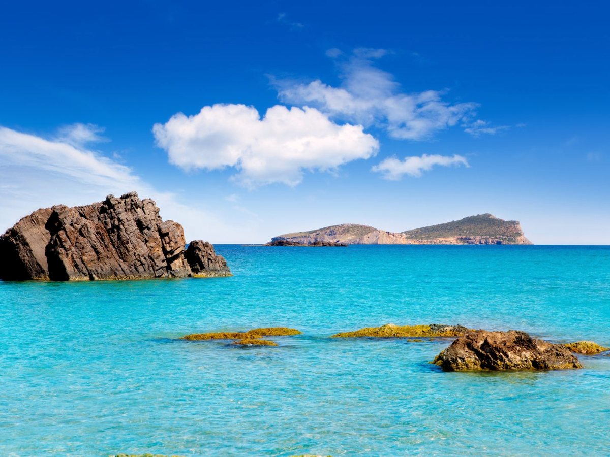 Bright turquoise shallows over scattered rocks with a rugged islet on the horizon under a blue, lightly clouded sky.