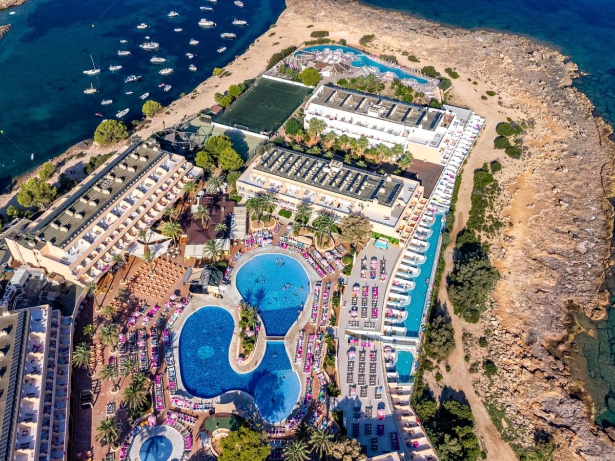Near-vertical aerial of the whole resort: two figure-eight pools, rows of loungers, a football pitch and low-rise blocks on a rocky headland beside a boat-filled bay.