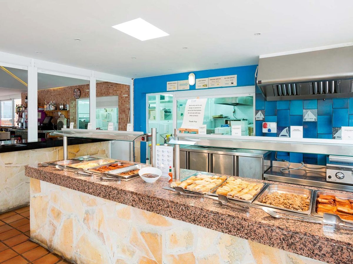 The buffet servery with trays of hot food along a stone-fronted counter, an open kitchen and a service-times sign behind.