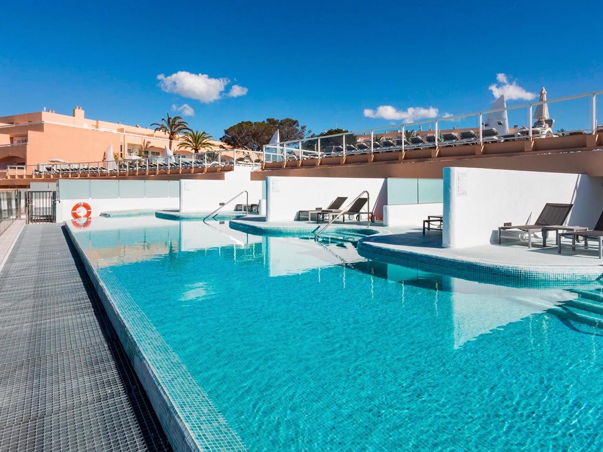 A turquoise pool with a level overflow edge, white-tiled terraces and a few loungers, the resort rising behind under a blue sky.