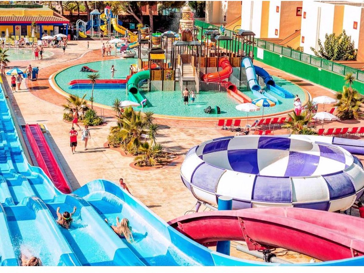 Blue and red flume slides and a large blue-and-white bowl slide beside the pirate-themed splash area, with children riding down and the resort behind.
