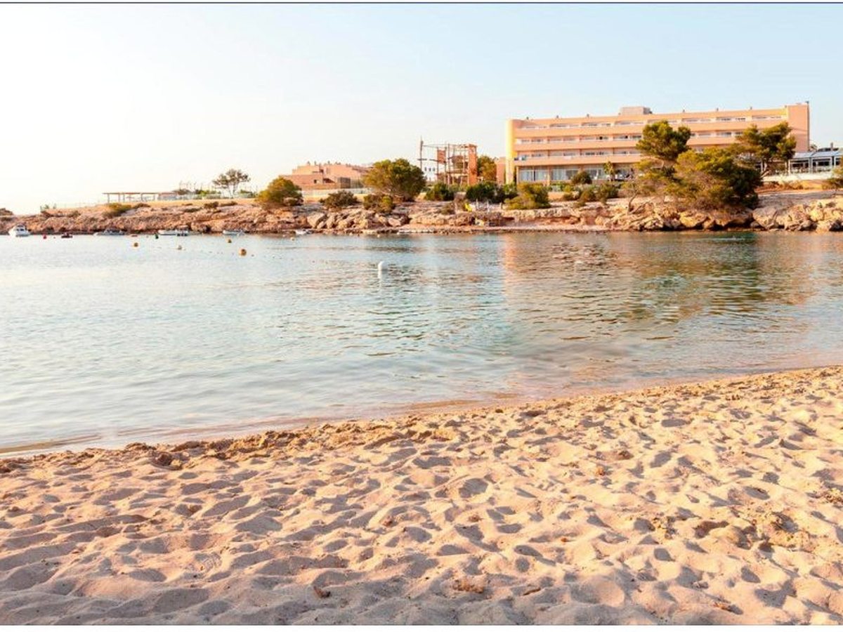 The sandy beach at Port d'es Torrent with calm water, moored boats and the resort on the low headland behind in warm evening light.