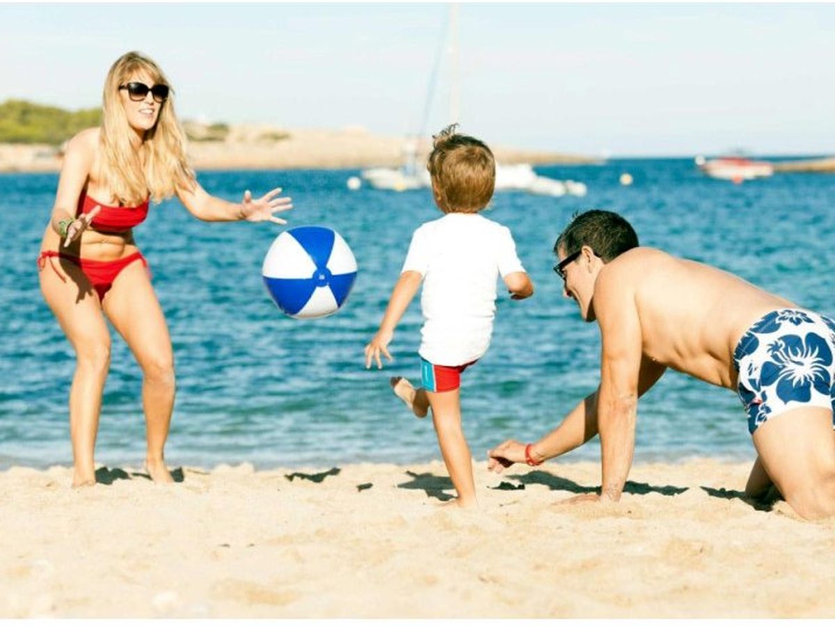 A parent and two children playing with a ball at the water's edge on the sandy beach, the sheltered bay and headland behind.
