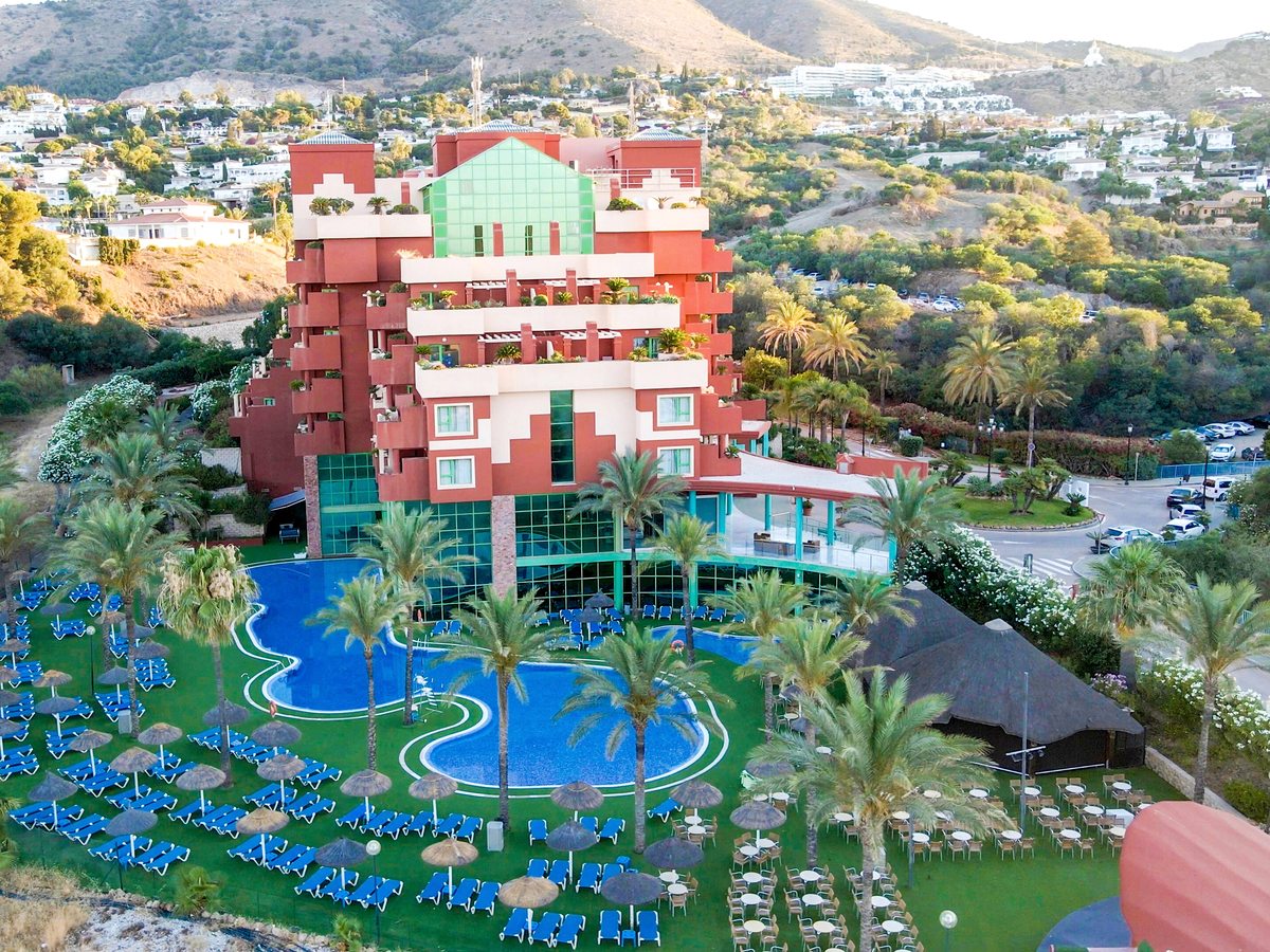 Aerial view of the red stepped hotel building above a winding blue pool ringed by palms, thatched parasols and rows of blue sun loungers, with hillside houses behind.