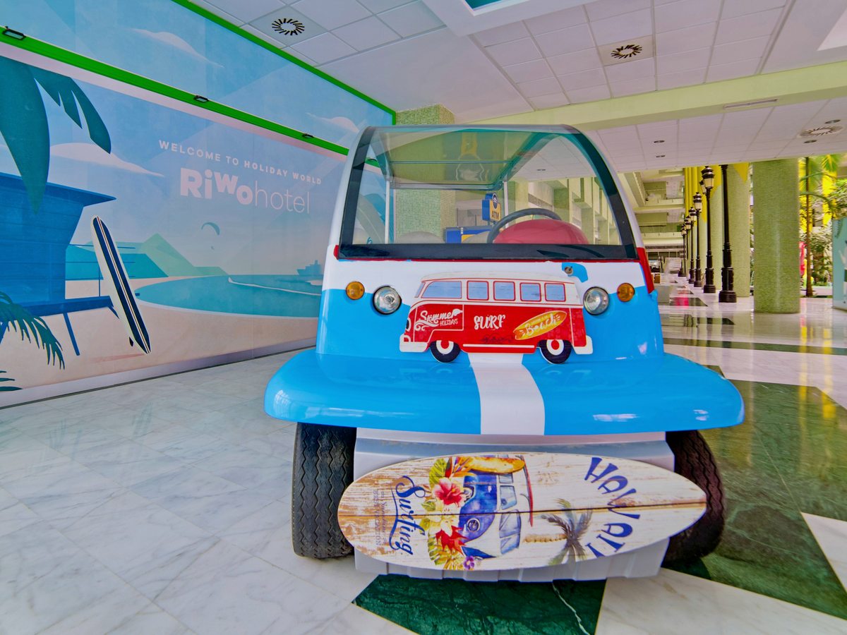 A blue-and-white surf-styled buggy in the lobby in front of a beach mural reading 'Welcome to Holiday World Riwo hotel', with a painted surfboard leaning against it.