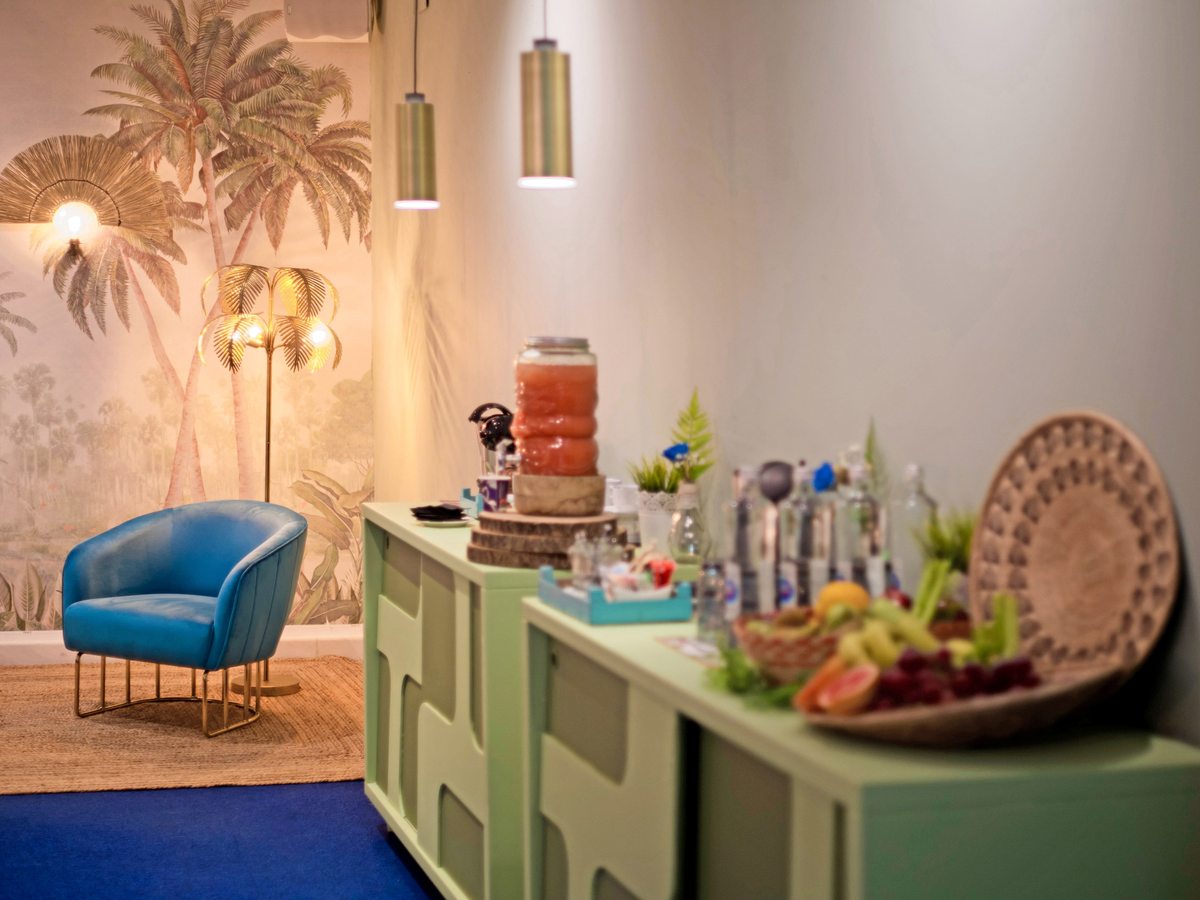 A corner of the Select lounge with a blue velvet armchair, a gold palm-frond floor lamp and a green sideboard set with drinks and a platter of fresh fruit against a painted palm mural.