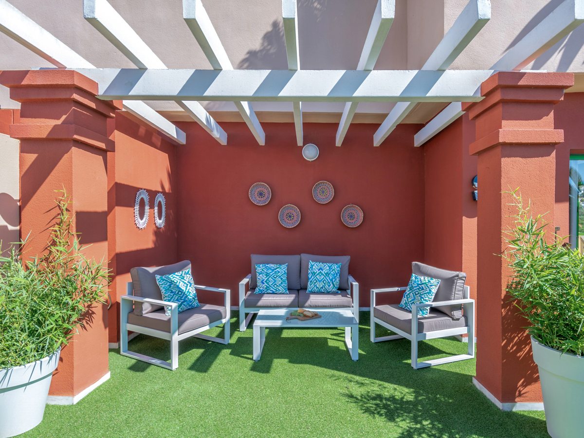 An enclosed courtyard with a white slatted pergola over grey-cushioned outdoor sofas and armchairs, potted bamboo and decorative woven discs on a terracotta wall.