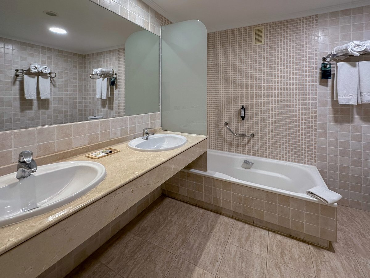 A bathroom with twin basins on a long vanity, a large mirror, a bathtub with a grab rail and glass screen, and towels on wall racks.