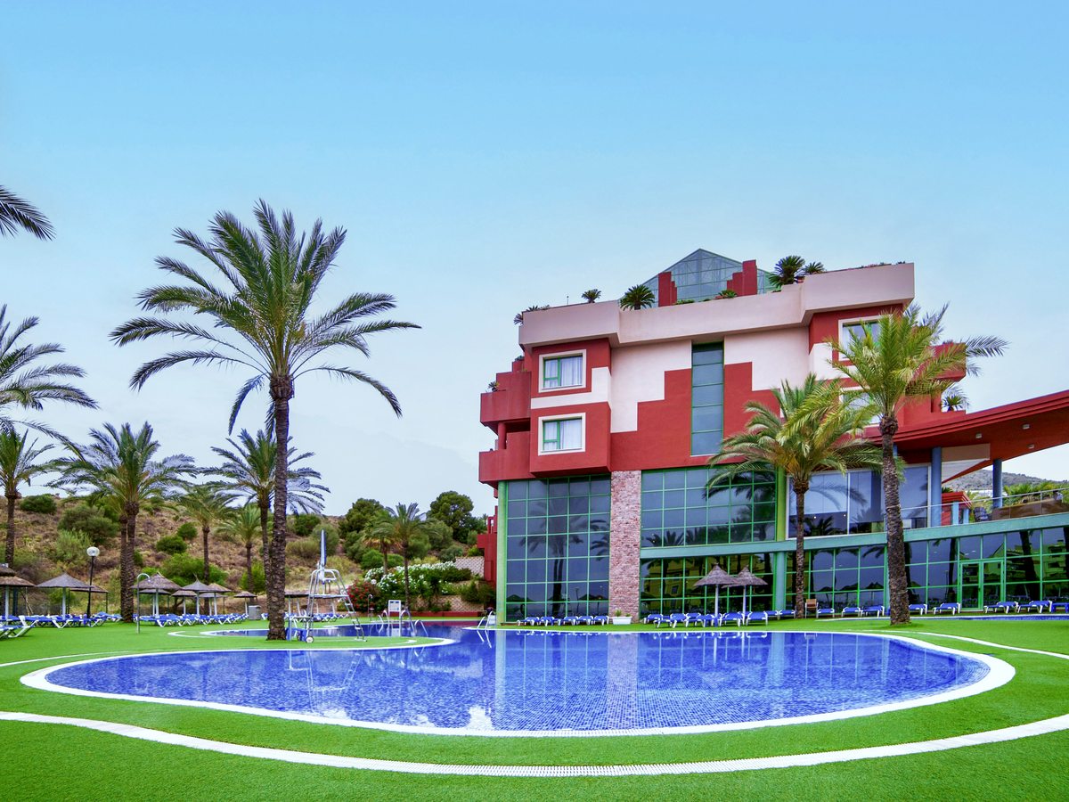 A curved outdoor pool on bright artificial lawn in front of the red-and-cream hotel building, edged with sun loungers, thatched parasols and palms under a clear sky.