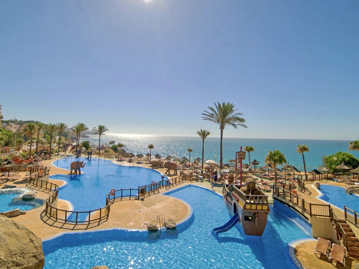 The Beach Club water park above the sea, with curved pools, a wooden pirate-ship play feature and small slide, elephant sculptures and thatched parasols.
