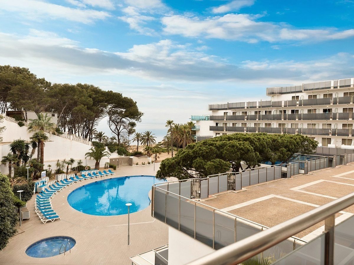 The sea-facing main pool with rows of blue loungers below the hotel's balconied building, pine trees and the sea beyond.