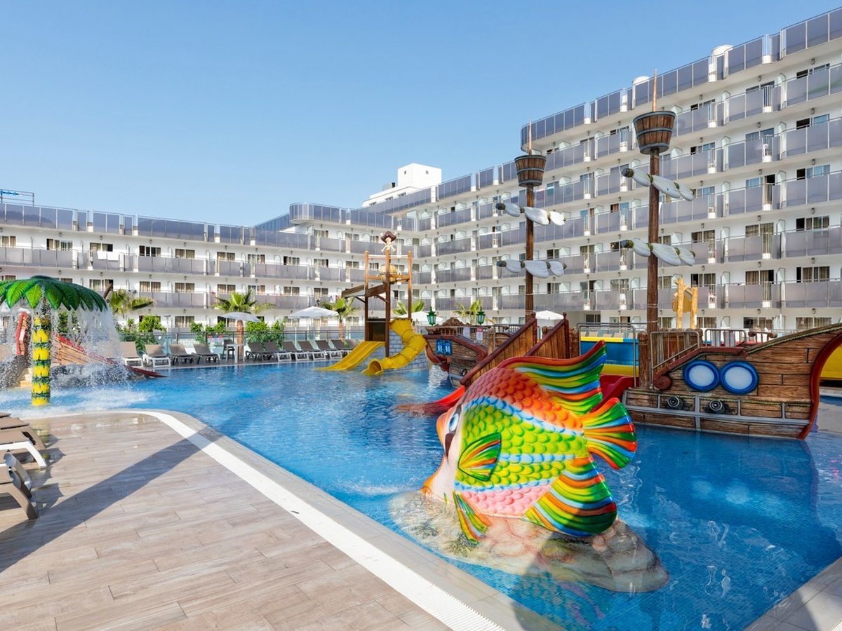 A shallow water-play area with a large colourful fish figure and gentle jets, the pirate ship beyond.