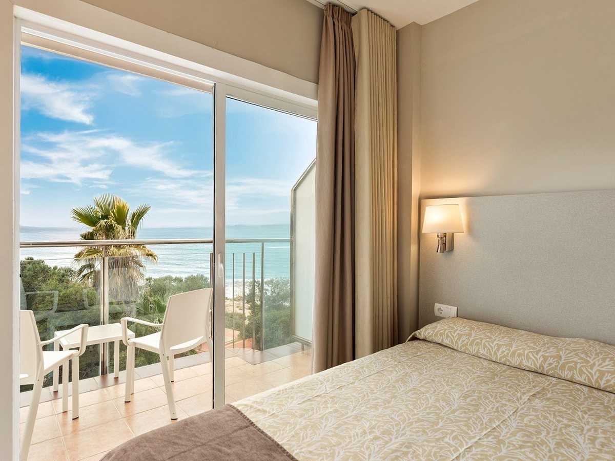 A room with a large bed and a glass-door terrace looking out over palms to the sea.