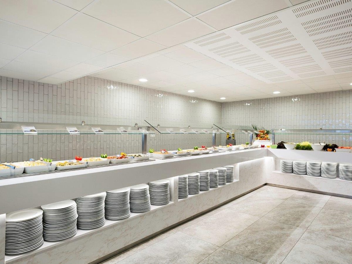 A long buffet serving counter with trays of salads and cold dishes behind glass guards, and tall stacks of plates on a shelf below.
