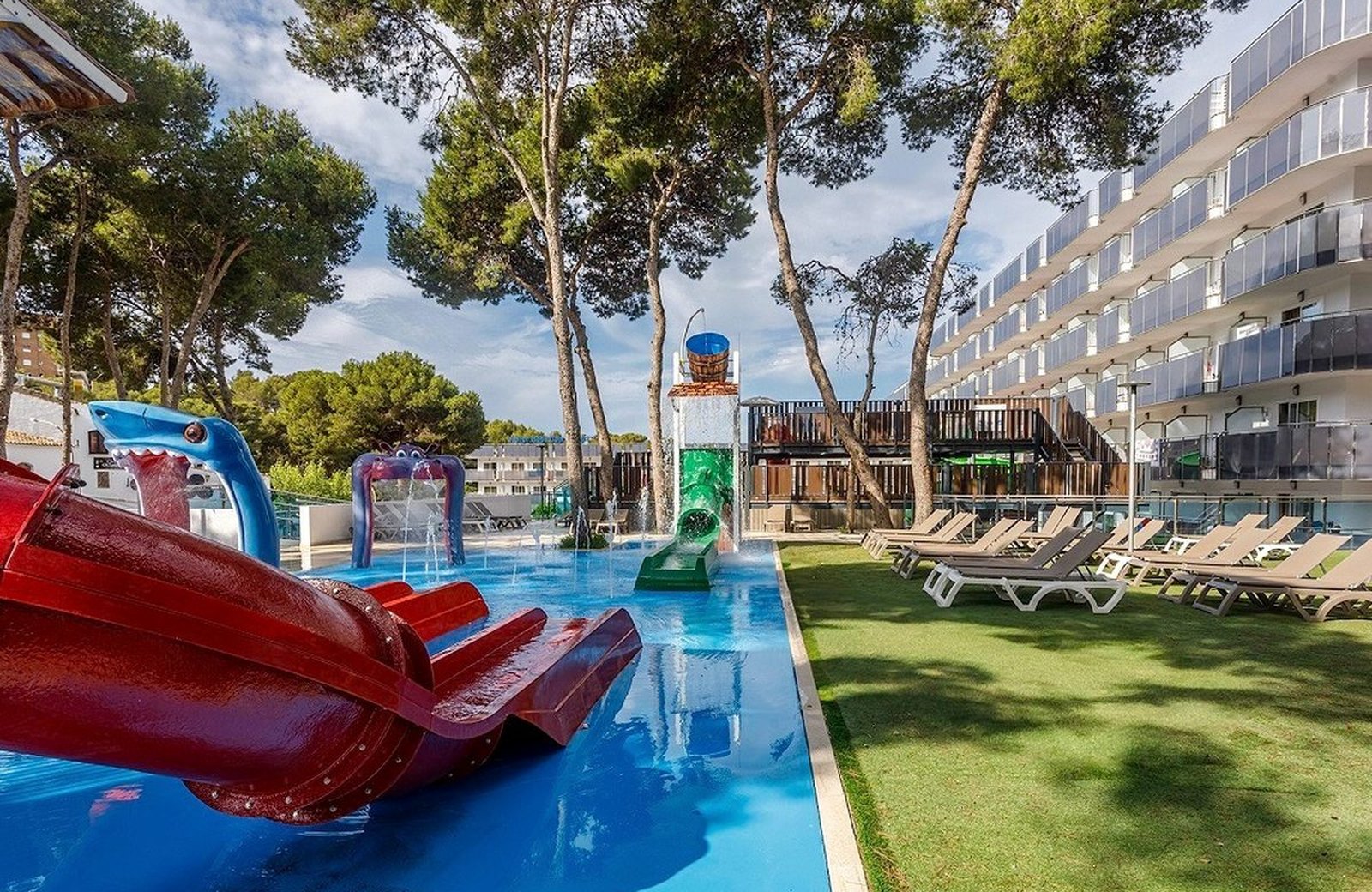 The children's water-play area of the splash pool at Hotel Best Cap Salou, with red tube slides, animal water-jet figures and a tipping-bucket tower beside a lawn of sun loungers, below the hotel's balconied building among pines.