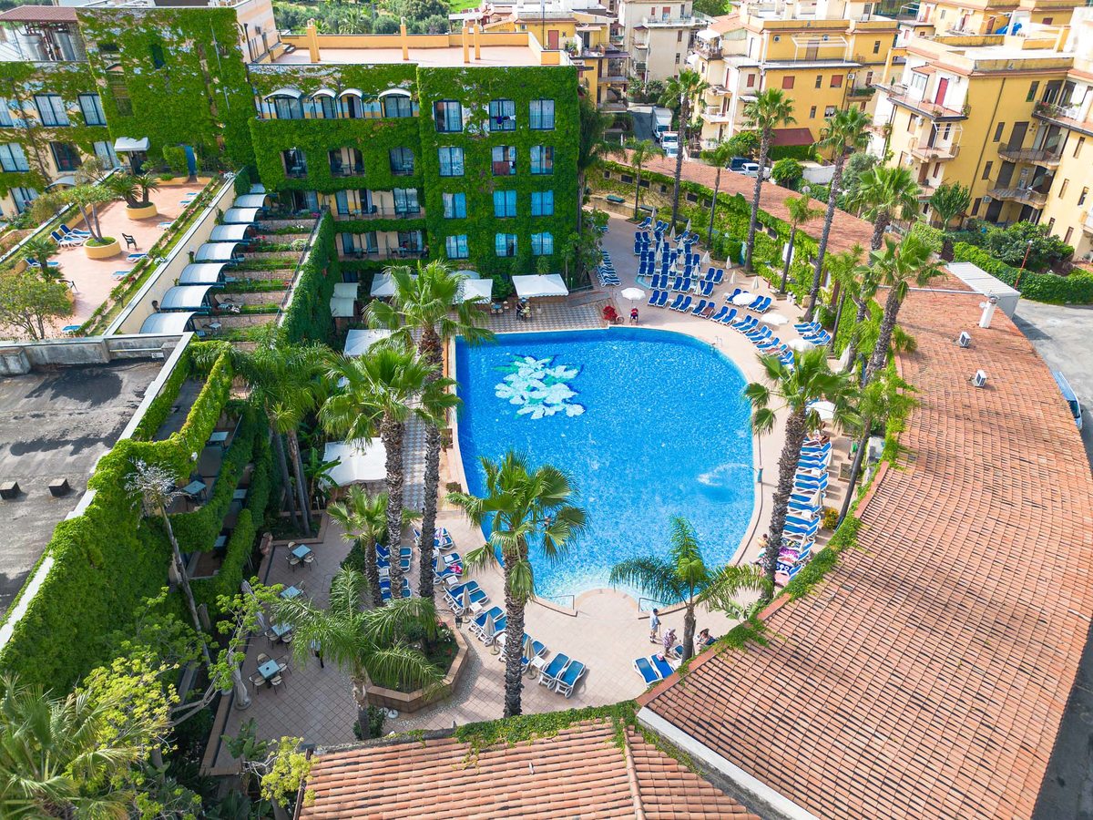 Aerial view of the ivy-covered hotel building enclosing a curved blue pool on a terracotta terrace, with palms, blue sun loungers and the town of Giardini Naxos around it.