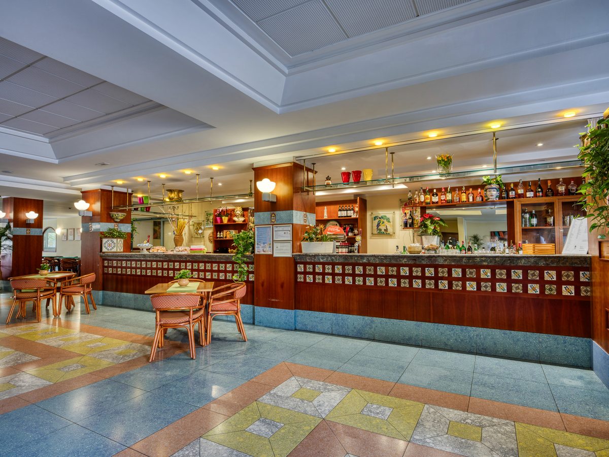 A long wooden and tiled bar counter with spirits on the shelves, pendant lights and rattan tables and chairs in front, on a patterned terrazzo floor.