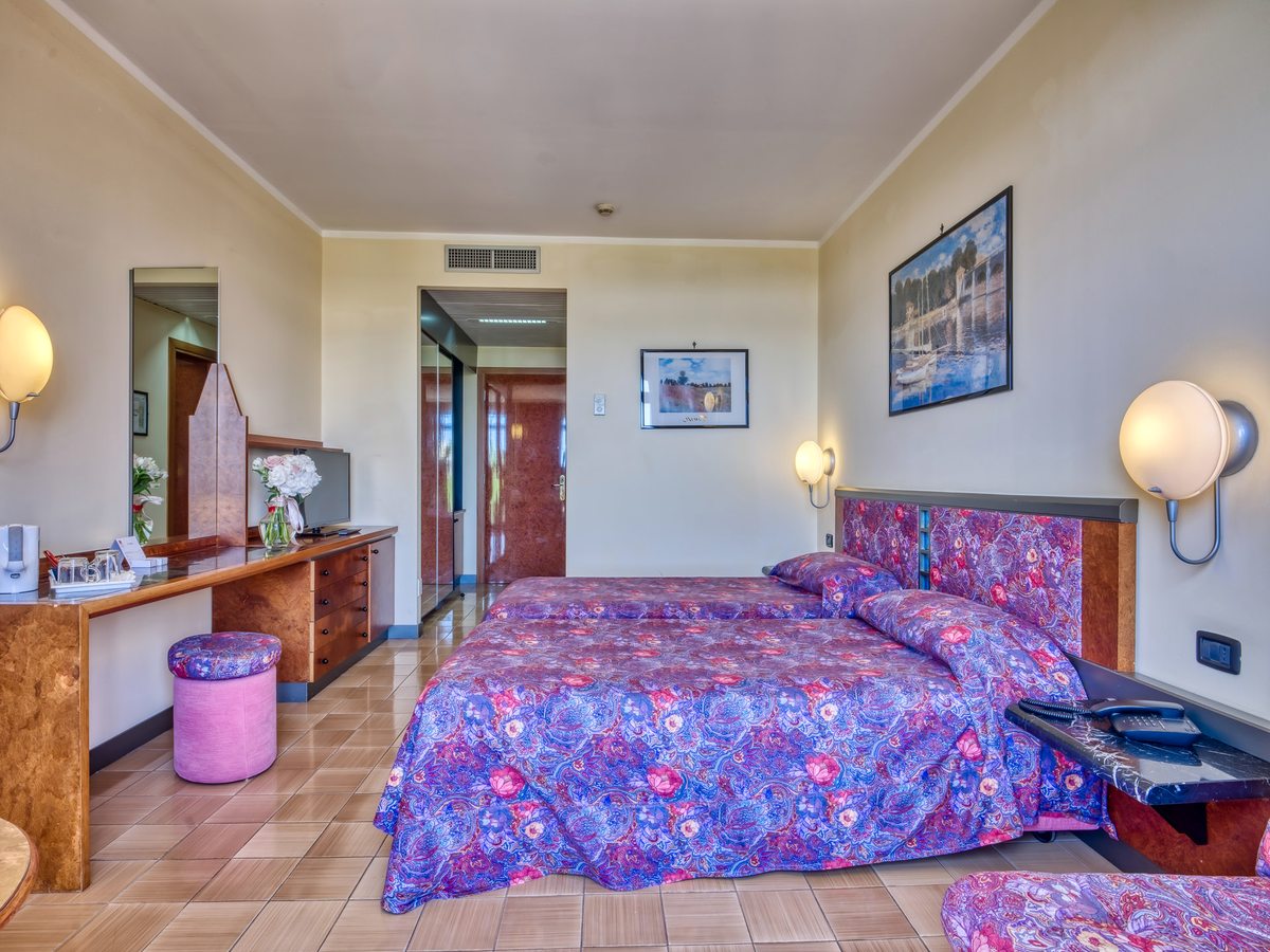 A hotel double room with a purple floral bedspread, a dark wood headboard, framed prints and wall lights, and a dressing table with a mirror opposite.